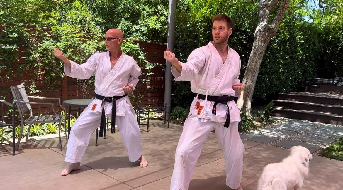 Eran Shine and his son Ori, both in white karate gi and black belts, holding a stance in a sunlit garden.