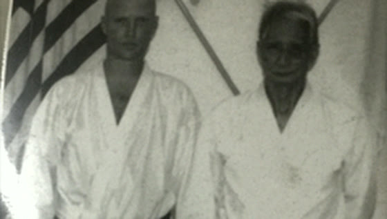 Eran Shine standing beside Sensei Hidetaka Nishiyama, both in white karate gi, in front of an American flag.